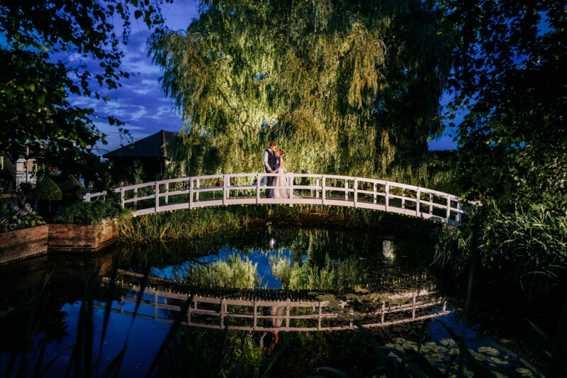 High House Weddings 1 A couple stood together in the middle of the bridge at High House at nighttime.