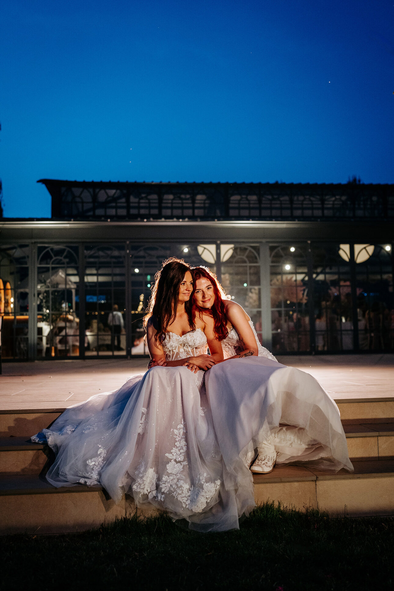 two brides sitting on the steps to the orangery at night cuddling up to each other. 