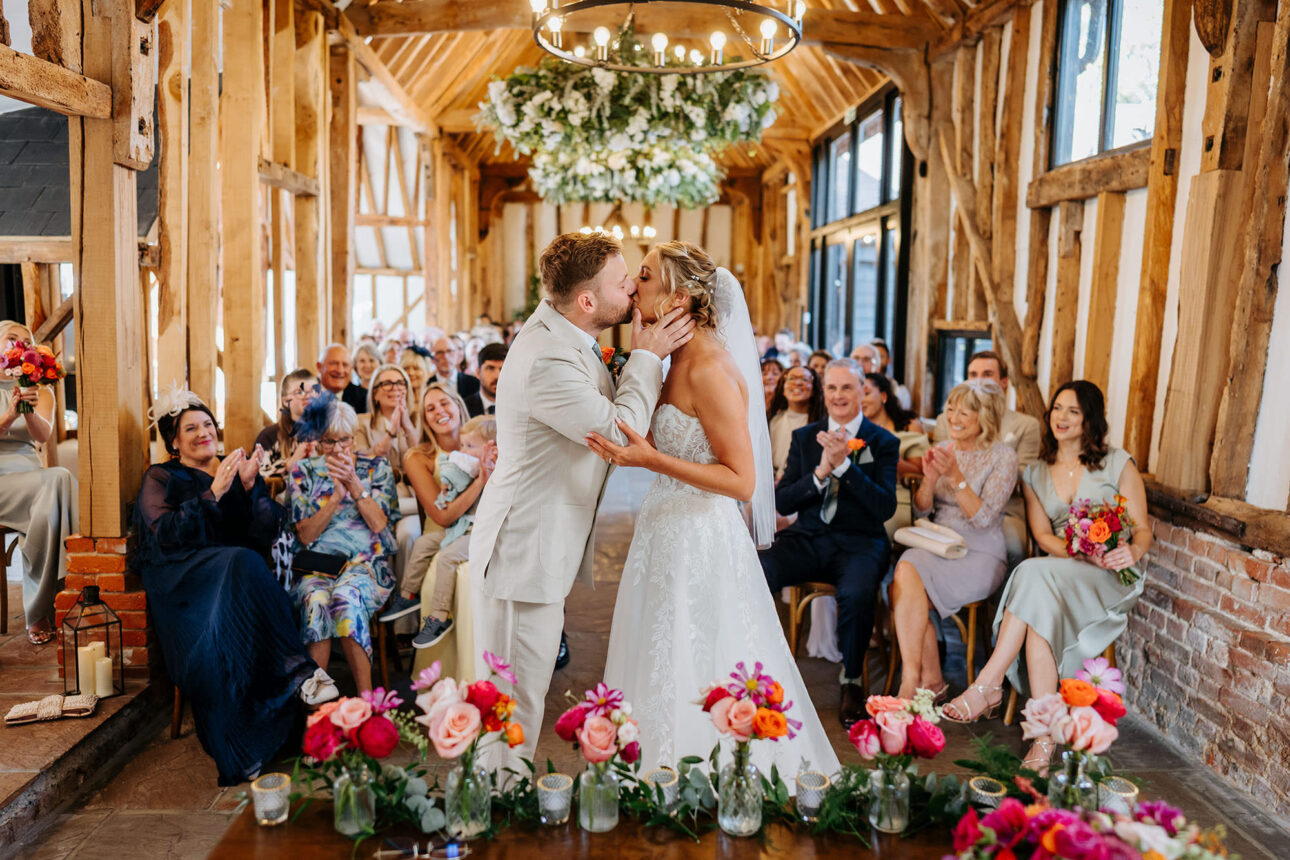 Bride & Groom having their first kiss at the ned of their wedding ceremony at the barn at High House, whilst their guests clap in the background. 